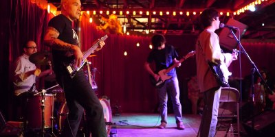 Band performing onstage with bass, guitars, and drums under warm lights in Austin, Texas.