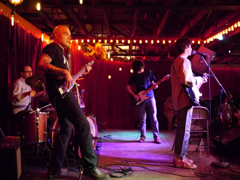 Band performing onstage with bass, guitars, and drums under warm lights in Austin, Texas.
